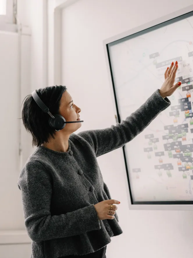 Junge Frau mit Headset steht vor einem Bildschirm, auf dem eine Software zu sehen ist, welche Taxis trackt. Sie berührt den Bildschirm mit ihrer Hand.