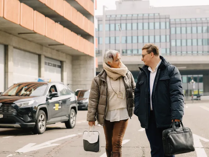 Draussen zwei Personen laufen zusammen, im Hintergrund ein Taxi und städtische Bauten